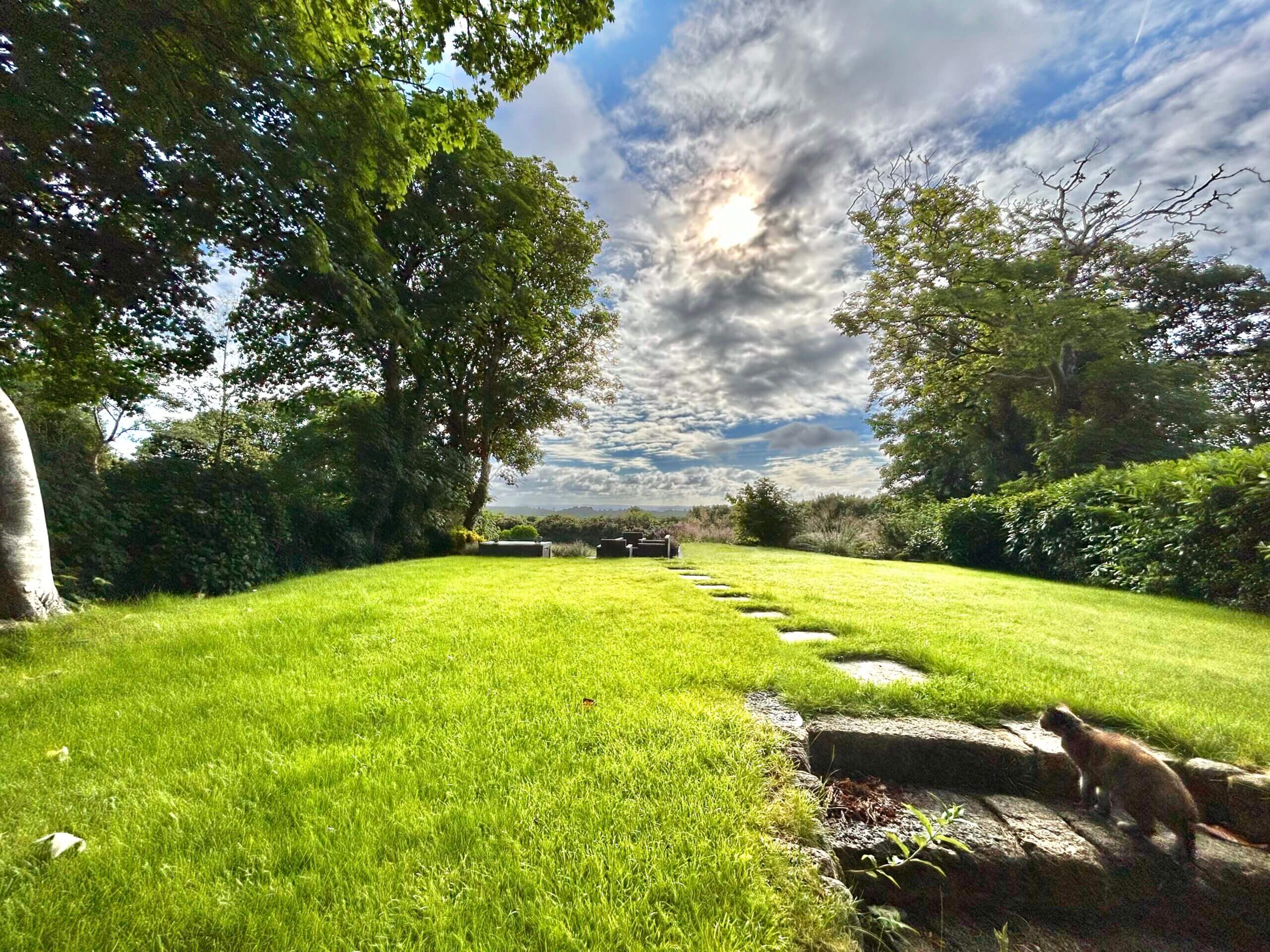 A lush green lawn with stone stepping paths winds through stunning private gardens, leading to benches under a partly cloudy sky. A cat stands near stone steps in the foreground, surrounded by trees and hedges. Sancreed House Retreat
