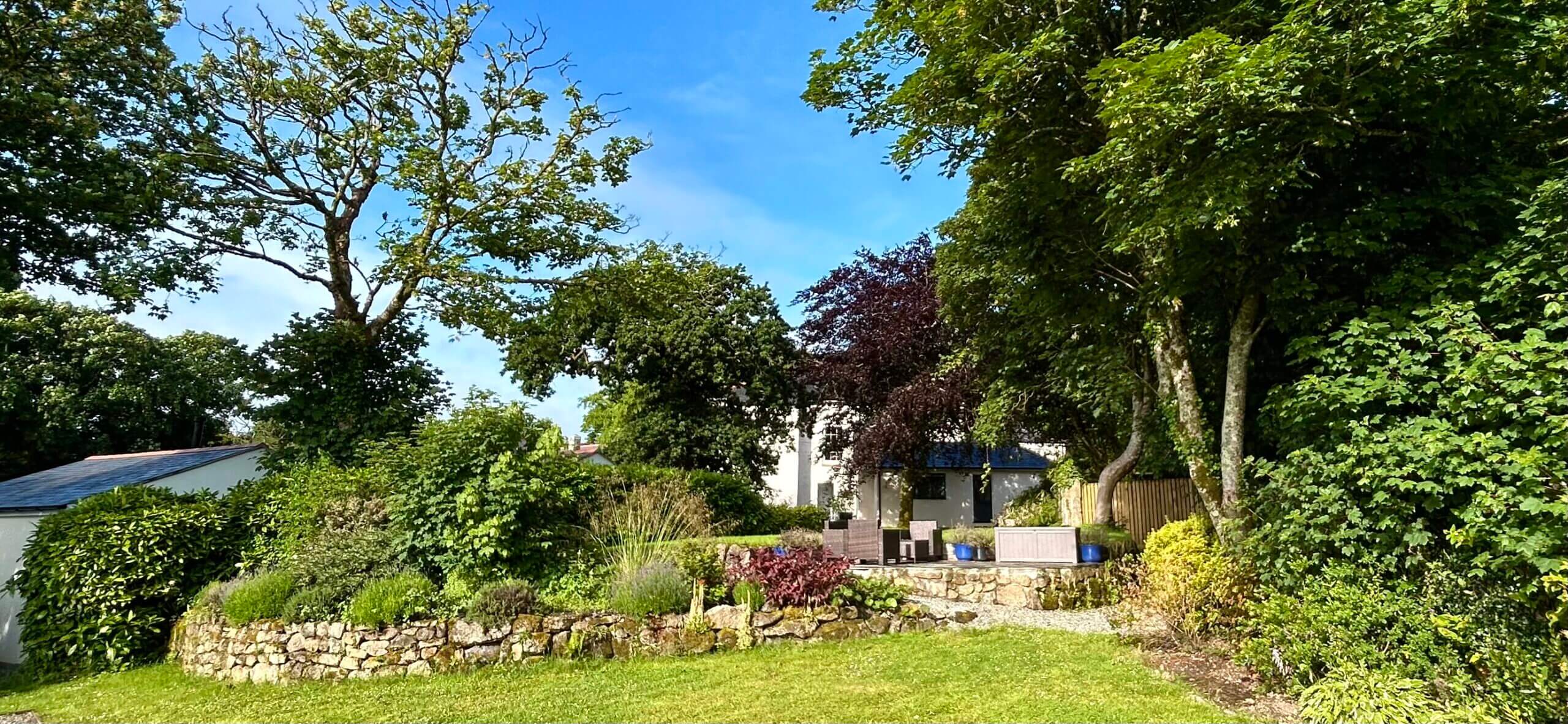 A lush garden with green grass, a stone wall, and various trees surrounds a white house partially visible in the background under a bright blue sky, showcasing stunning private gardens. Sancreed House Retreat