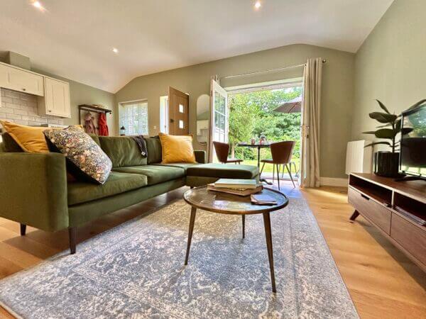 A cozy living room with a green sofa, patterned cushions, a round coffee table, and a rug. Glass doors open to a patio with outdoor seating and greenery. Enjoy this inviting space in one of our Luxury Cottages Penzance for the perfect retreat. Sancreed House Retreat