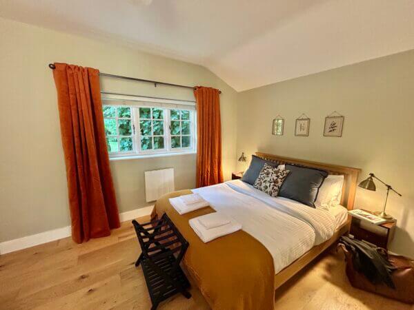 Cozy bedroom in one of our Premium self-catering cottages, featuring a wooden bed, mustard throw, white bedding, blue pillows, and inviting orange curtains. Nightstands with lamps and tasteful decor complete this Luxury Cottage Penzance experience. Sancreed House Retreat