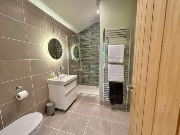 Modern bathroom in one of our luxury cottages Penzance, featuring beige tiles, a round backlit mirror above a white sink cabinet, glass-enclosed shower with gray tiles, wall-mounted towel rack, and a stylish wooden door. Sancreed House Retreat
