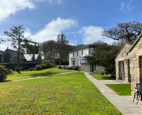 A well-maintained grassy lawn with stone paths leads to a large white house with stone accents, surrounded by trees and premium self-catering cottages under a partly cloudy blue sky. Sancreed House Retreat