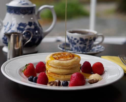 A stack of pancakes is topped with syrup and surrounded by strawberries, raspberries, blueberries, pecans, and a dollop of butter. In the background, there is a teapot, a cup of tea, and a small pitcher. Sancreed House Retreat
