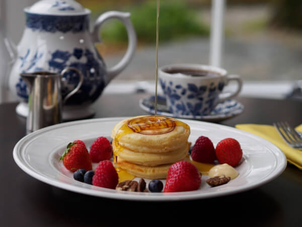 A stack of pancakes is topped with syrup and surrounded by strawberries, raspberries, blueberries, pecans, and a dollop of butter. In the background, there is a teapot, a cup of tea, and a small pitcher. Sancreed House Retreat