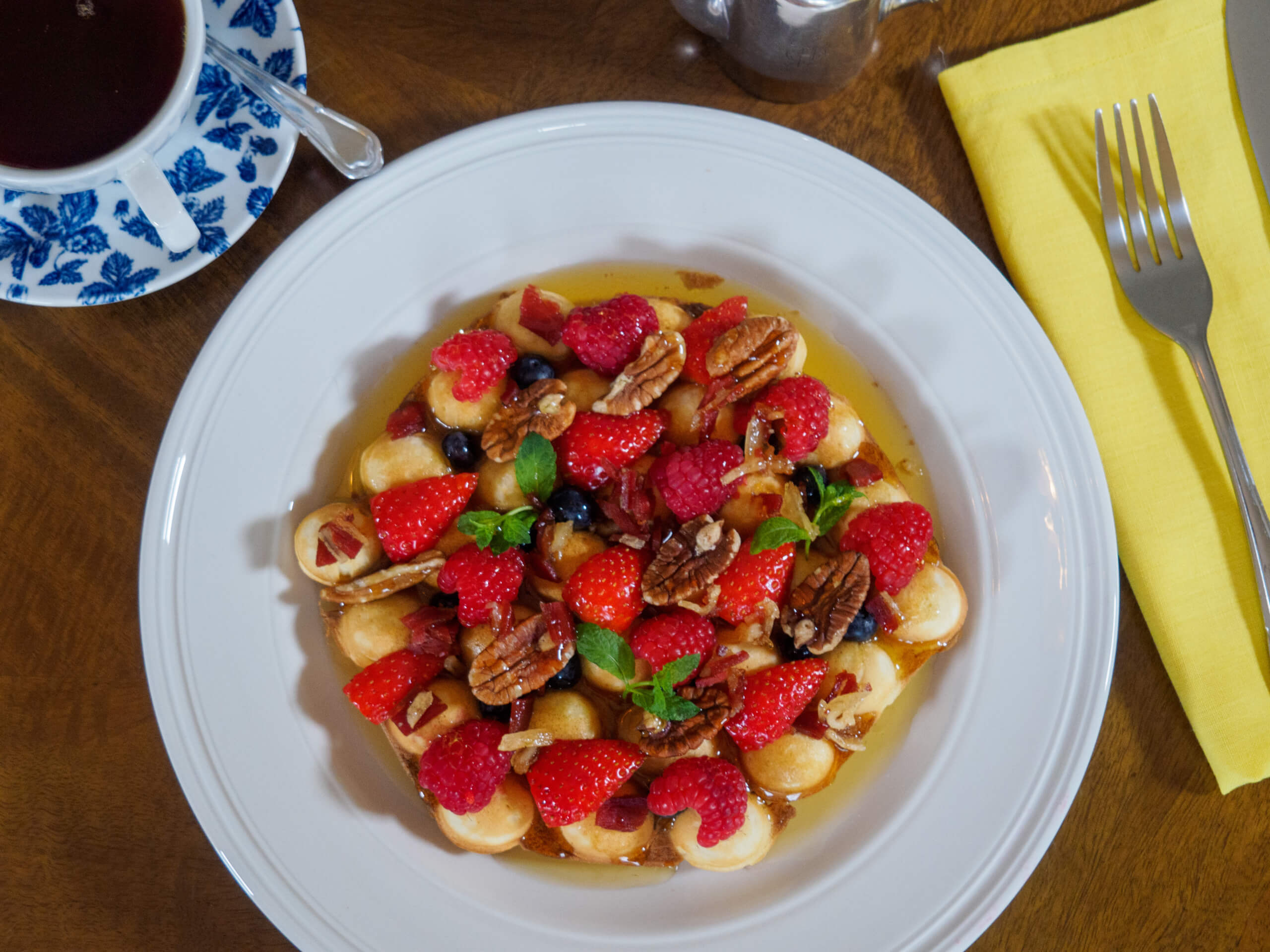 A white plate with mini pancakes topped with raspberries, strawberries, blueberries, pecans, and mint, drizzled with syrup. A cup of tea, a yellow napkin, fork, and teapot are nearby on a wooden table. Sancreed House Retreat