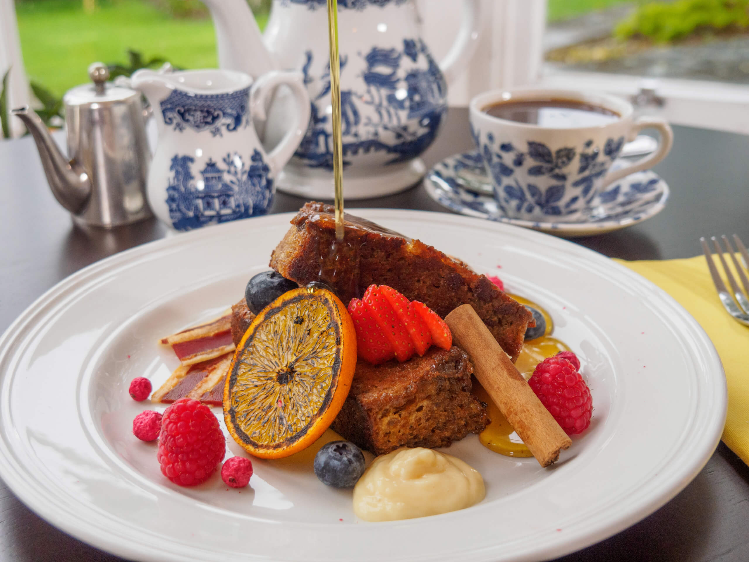 A plate of French toast topped with fresh berries, orange slice, cinnamon stick, and sauce is being drizzled with syrup. In the background are a teapot, cup of coffee, and blue-and-white china. Sancreed House Retreat