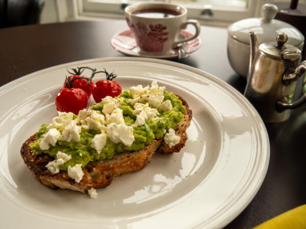 A plate with smashed avocado and crumbled feta on toast, served with roasted cherry tomatoes. In the background, a teacup, a teapot, and a metal milk jug sit on a dark table near a window. Sancreed House Retreat