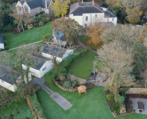 Aerial view of a landscaped property with multiple white buildings, green lawns, trees, a small gazebo, pathways, and a fenced area with lounge chairs, all surrounded by stunning private gardens and lush foliage. Sancreed House Retreat