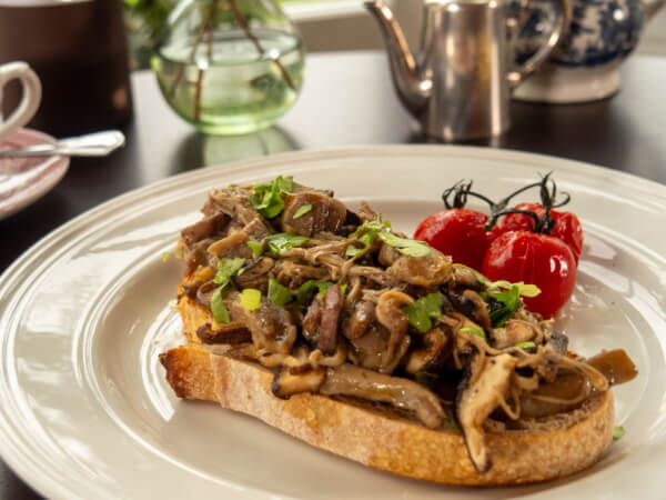 A slice of toasted bread topped with sautéed mushrooms and herbs is served on a white plate, with roasted cherry tomatoes on the side. A teapot and glass vase are blurred in the background. Sancreed House Retreat