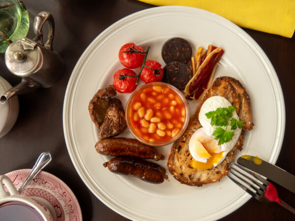 A plate of full English breakfast with poached eggs on toast, baked beans, sausages, bacon, grilled tomato, black pudding, and mushrooms, set on a dark table with tea and a yellow napkin nearby. Sancreed House Retreat