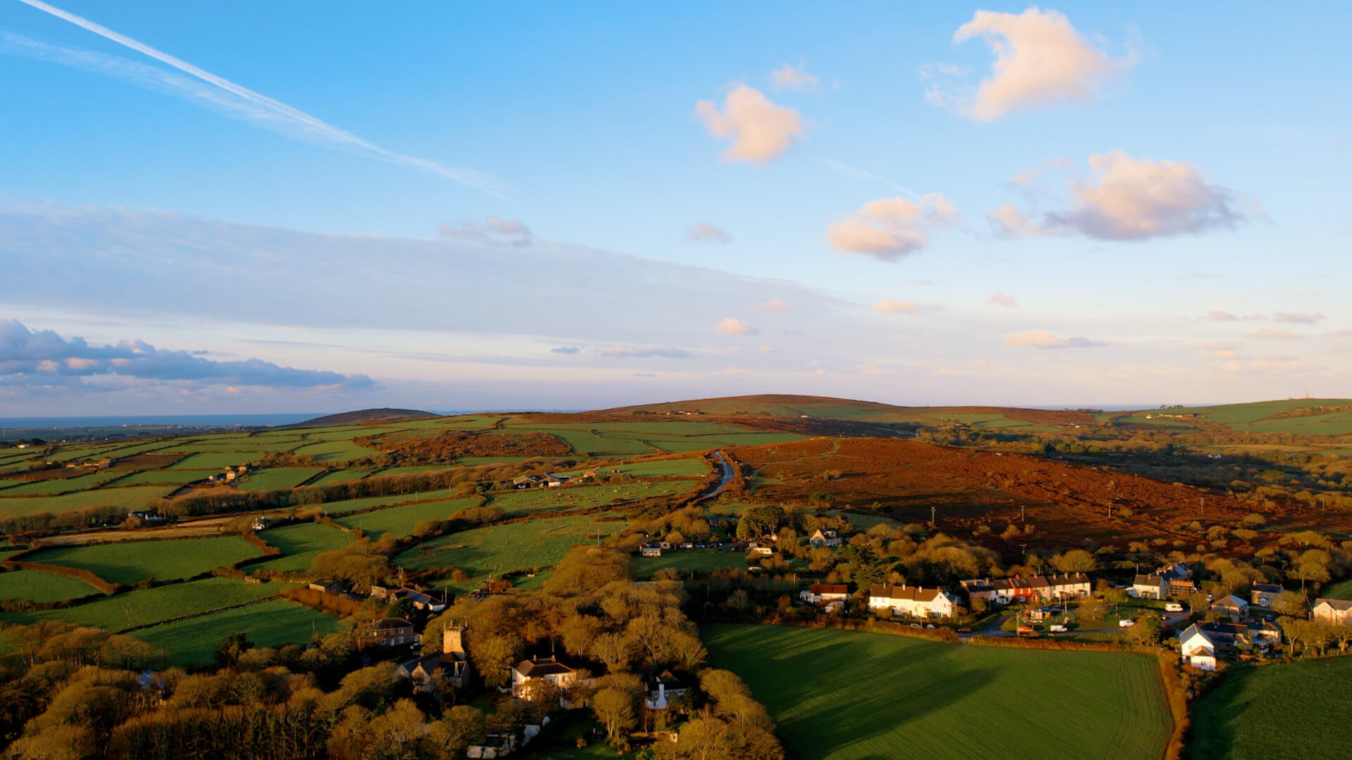 Aerial view of a rural landscape with green fields, scattered houses, and winding roads—perfect for exploring local things to do—set against hills and a blue sky with fluffy clouds at sunset. Sancreed House Retreat
