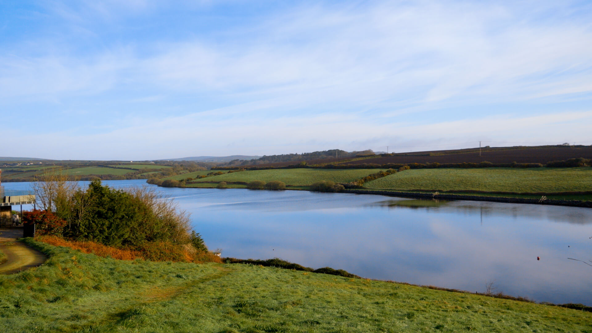 A serene lake bordered by green fields and rolling hills under a blue sky with light clouds; among the picturesque scenery, discover relaxing things to do near the trees and small building at the water’s edge in the foreground. Sancreed House Retreat