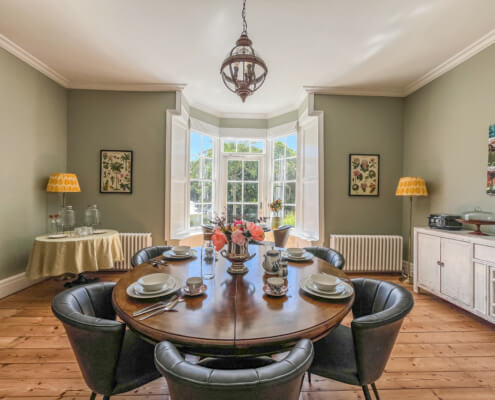 A bright dining room at this inviting Bed and Breakfast features a round wooden table set for six, black chairs, and a vase of flowers. Sunlight pours through bay windows, with yellow lamps, framed art, and a buffet cabinet adding charm. Sancreed House Retreat