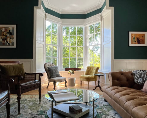 A cozy living room with dark green walls, a large bay window letting in sunlight, and various chairs and a brown tufted sofa around a glass coffee table. There are lamps and framed art on the walls. Sancreed House Retreat