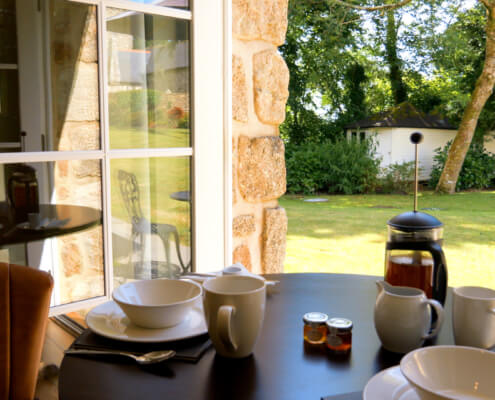 A table set for breakfast with bowls, cups, a French press, and small jars of jam near a window at a luxury retreat in Penzance, overlooking a sunny garden with green grass, trees, and a small white shed. Sancreed House Retreat