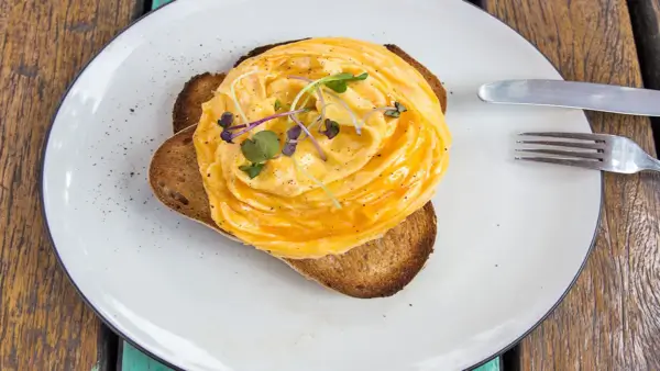 A plate with two slices of toasted bread topped with creamy scrambled eggs, garnished with microgreens. A fork and knife rest on the side of the plate, which sits on a rustic wooden table. Sancreed House Retreat