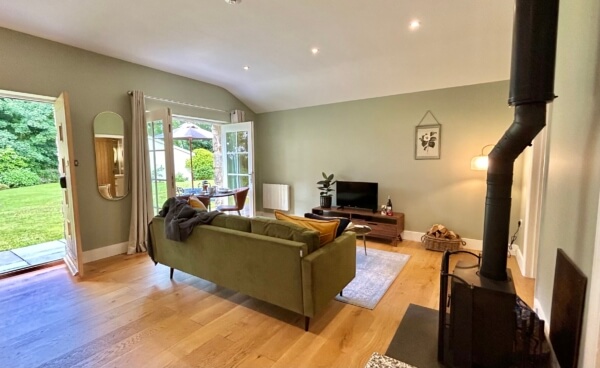 A cozy living room with light green walls, wood flooring, a green sofa, TV on a stand, and a wood-burning stove reflects the charm of luxury cottages in Penzance. Glass doors open to a lush garden, while art and mirrors create a warm atmosphere. Sancreed House Retreat