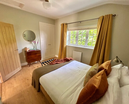 A cozy bedroom in one of our Luxury Cottages Penzance, featuring a neatly made double bed, orange pillows, mustard curtains, wooden floors, and a dresser with a round mirror—perfect for a premium self-catering cottage stay. Sancreed House Retreat