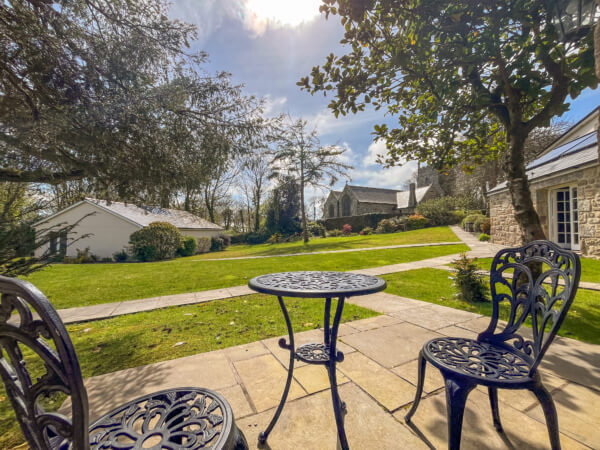A sunny garden patio with a round metal table and two ornate chairs overlooks the lawn and stone buildings of Luxury Cottages Penzance, offering a bright, pleasant day in premium self-catering cottages surrounded by greenery. Sancreed House Retreat