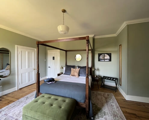 A cozy bedroom at our Bed & Breakfast in Penzance features sage green walls, a four-poster bed with gray and tan bedding, a green ottoman, wall mirrors, a small desk, wooden floors, and an area rug for added comfort. Sancreed House Retreat