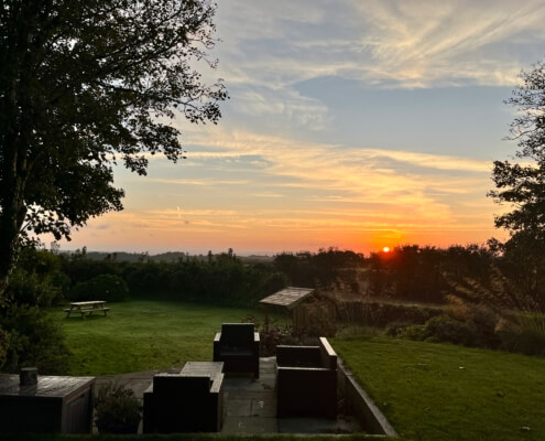 View of stunning private gardens at sunset with outdoor seating, a wooden bench on the lawn, and trees framing the scene. The sun sets near the horizon, casting an orange glow over the sky and landscape. Sancreed House Retreat