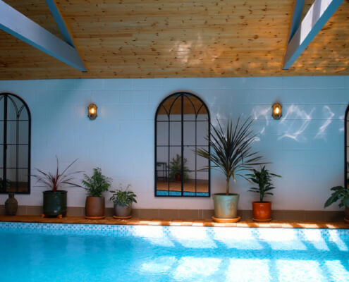 Indoor swimming pool with clear blue water, potted plants along the poolside, arched mirrors on a white wall, and a wooden ceiling with skylights—a true Luxury Retreat in Penzance. Sancreed House Retreat