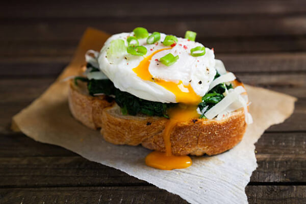 A slice of toasted bread topped with cooked spinach, shredded cheese, and a poached egg with a runny yolk, garnished with chopped green onions, sits on parchment paper on a wooden surface. Sancreed House Retreat