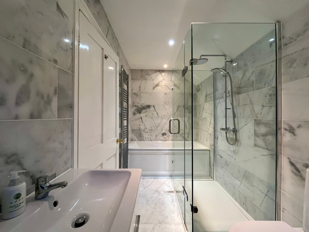 Modern bathroom with marble walls and floor at our Bed & Breakfast in Penzance, featuring a white sink, glass-enclosed shower, bathtub, and a chrome towel radiator, all under bright lighting. Sancreed House Retreat