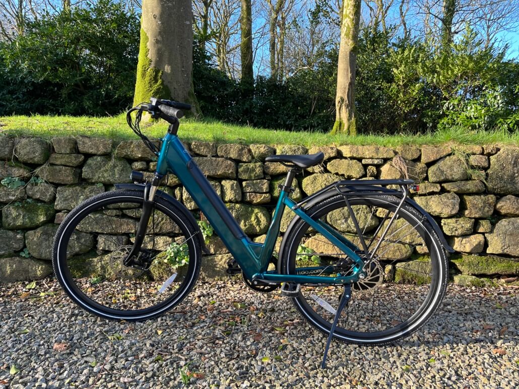 A blue electric bicycle with a step-through frame, available with additional services, is parked on gravel in front of a stone wall and green trees on a sunny day. Sancreed House Retreat