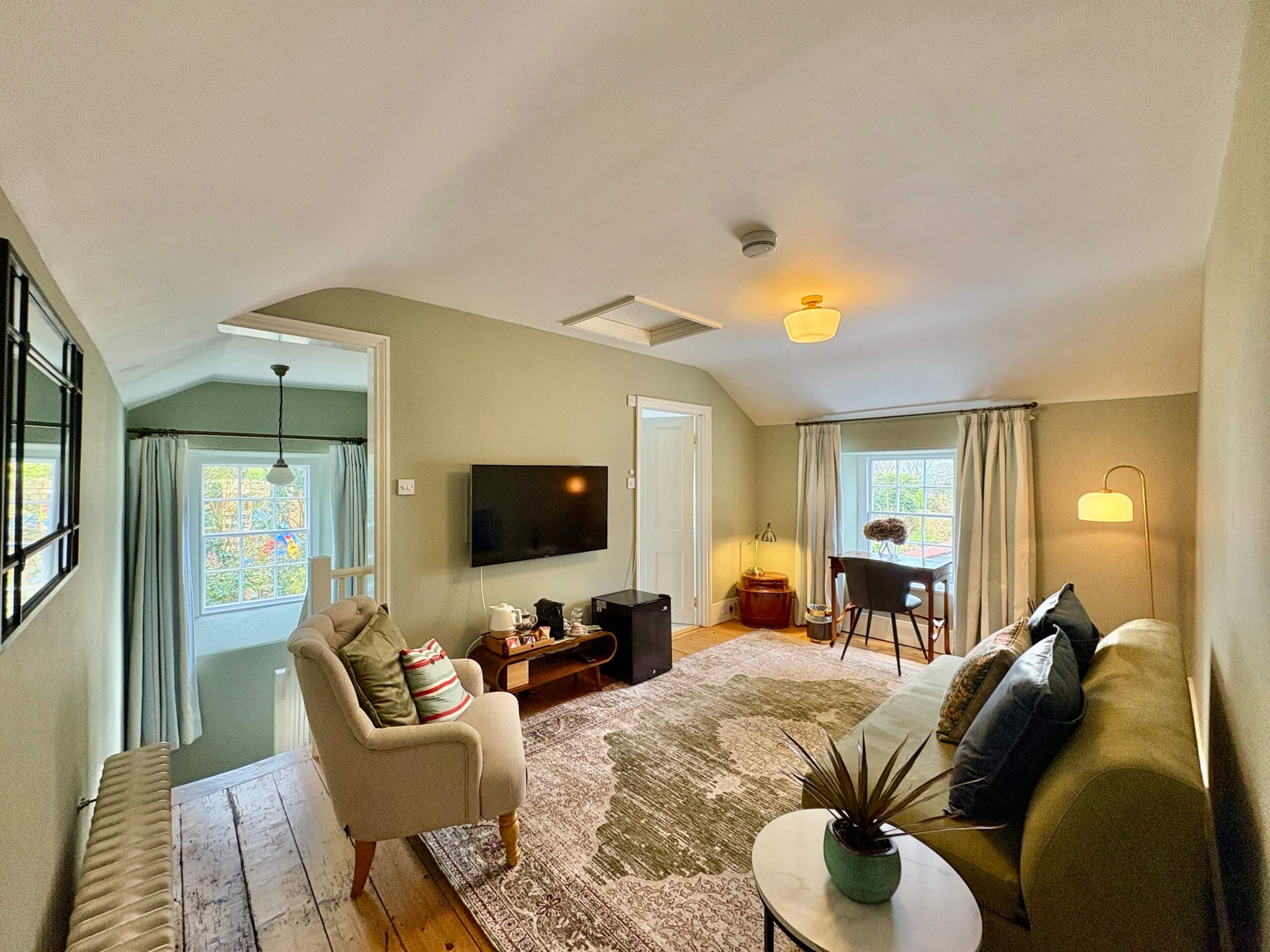 A cozy, light-filled living room at this Bed & Breakfast in Penzance features a green sofa, armchair, TV, and a small desk by the window. Wooden floors and neutral walls create a warm, inviting atmosphere for your stay. Sancreed House Retreat