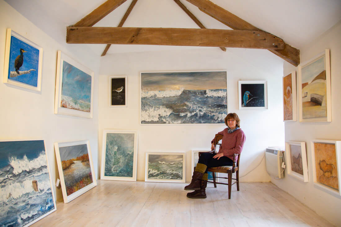A woman sits on a chair in a small art gallery with white walls, surrounded by framed paintings of seascapes, birds, and animals displayed on the walls and leaning on the floor. Exposed wooden beams are visible above. Sancreed House Retreat