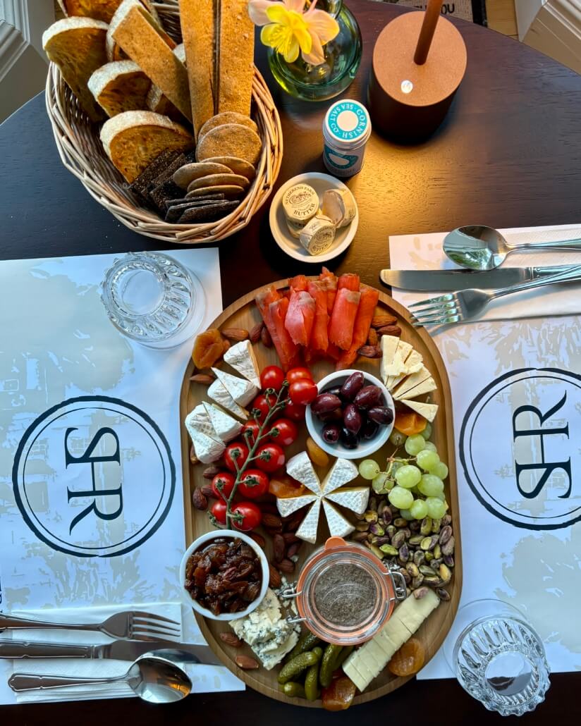 A wooden table set for two with placemats, cutlery, and napkins features a platter of cheese, cherry tomatoes, grapes, nuts, dried fruit, olives, smoked salmon, and pickles. Enjoy fresh bread and flowers—ask about our Additional Services for a tailored touch. Sancreed House Retreat