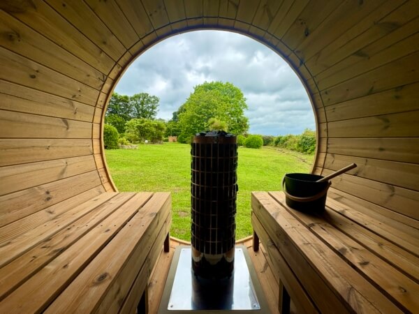 View from inside a wooden barrel sauna at a luxury retreat in Penzance, looking out onto a green grassy field. Benches line either side, with a central heater and a wooden bowl with ladle on the right. Trees and cloudy sky are visible outside. Sancreed House Retreat