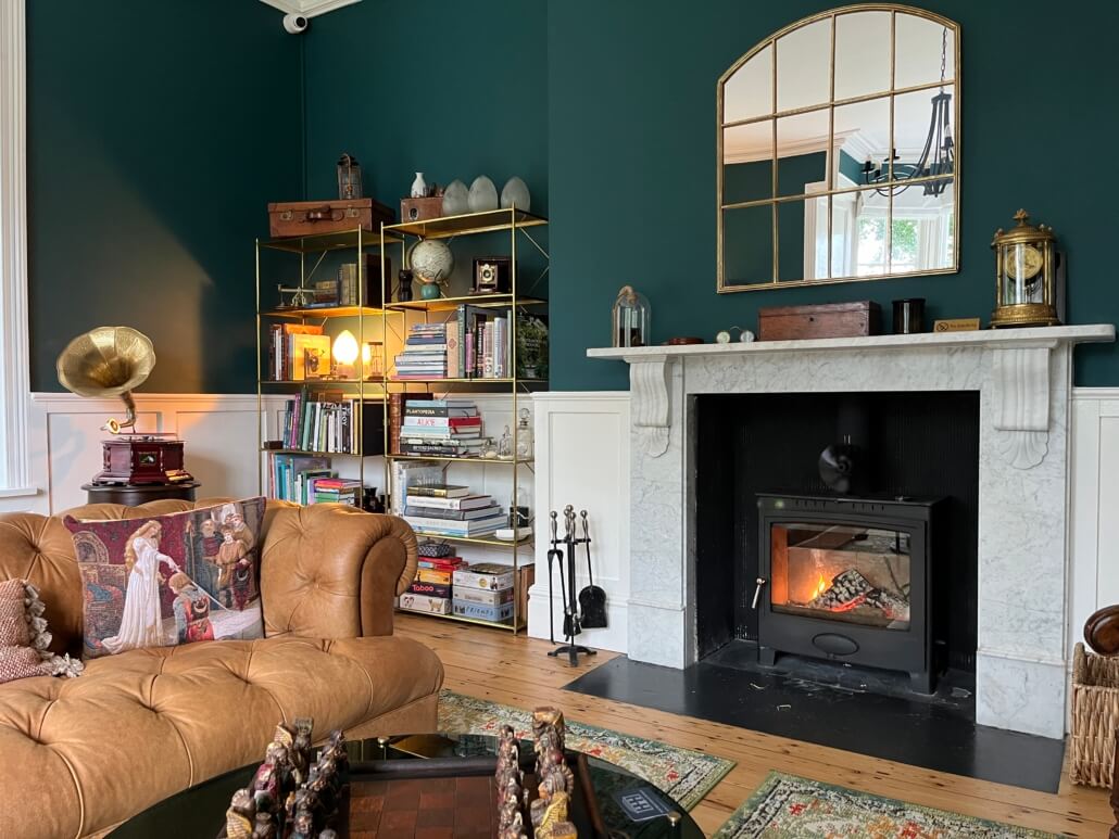 A cozy living room with dark green walls, a white marble fireplace with a fire burning, gold shelves filled with books and decor, a tan tufted sofa, and a chessboard on a table in the foreground. Sancreed House Retreat