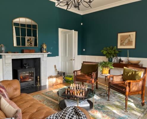 A cozy living room with dark green walls, a white fireplace, a brown leather sofa, two wooden chairs, plants, a chessboard on a glass coffee table, and natural light entering from large windows. Sancreed House Retreat