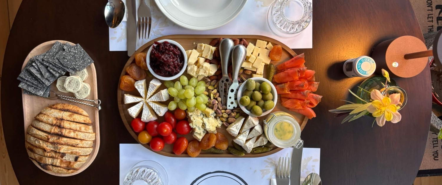 A round table set for two with plates, cutlery, and placemats. In the center is a charcuterie board with cheeses, meats, olives, tomatoes, and fruit. A small breadboard and crackers sit at the front, with a flower vase and lamp above. Sancreed House Retreat