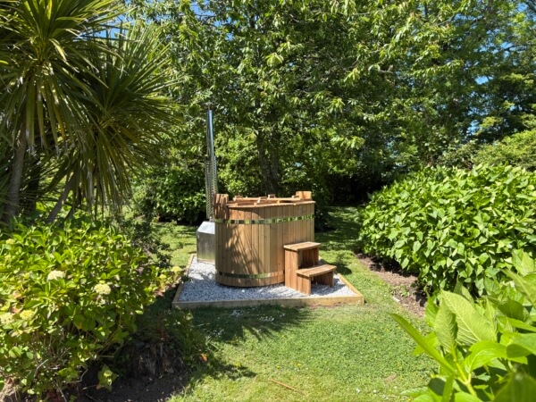 A wooden outdoor hot tub with a small set of steps sits on a gravel base surrounded by lush green grass, trees, and bushes on a sunny day. Sancreed House Retreat