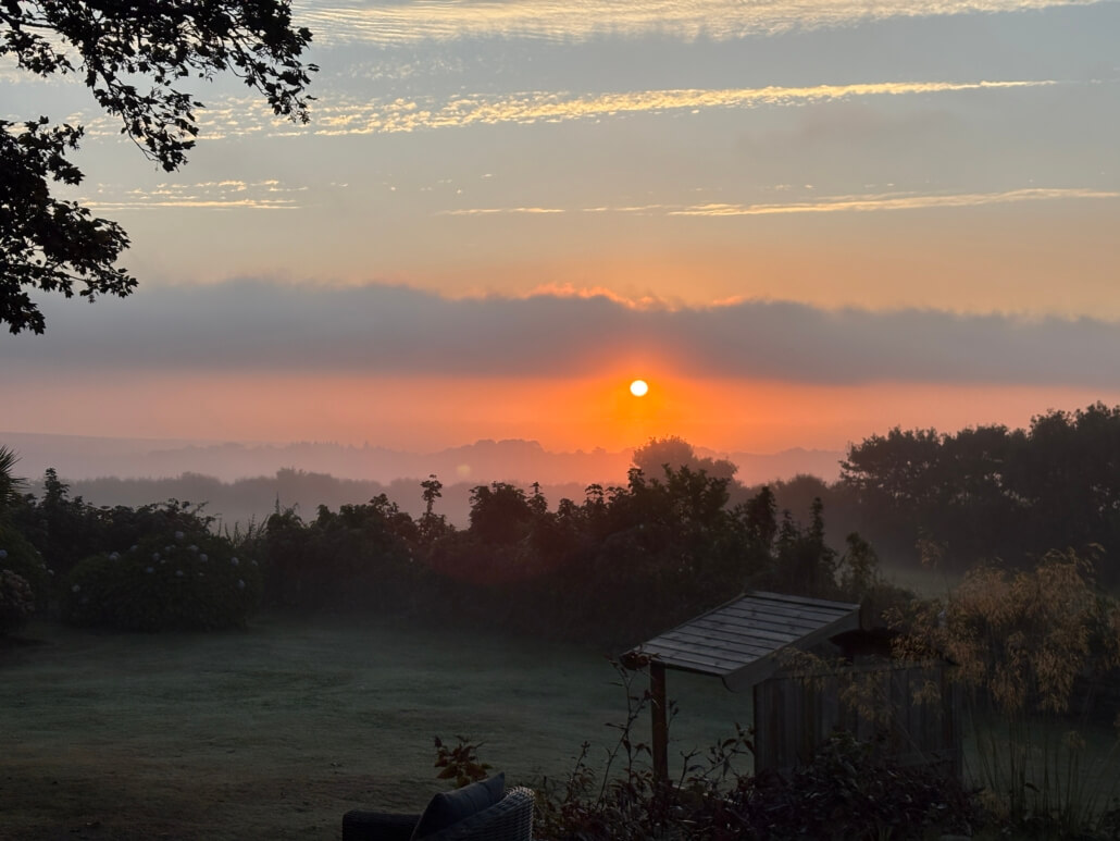 A sunrise over a misty landscape with trees and bushes in silhouette, an orange sun rising above the horizon, and a wooden garden structure in the foreground. Sancreed House Retreat