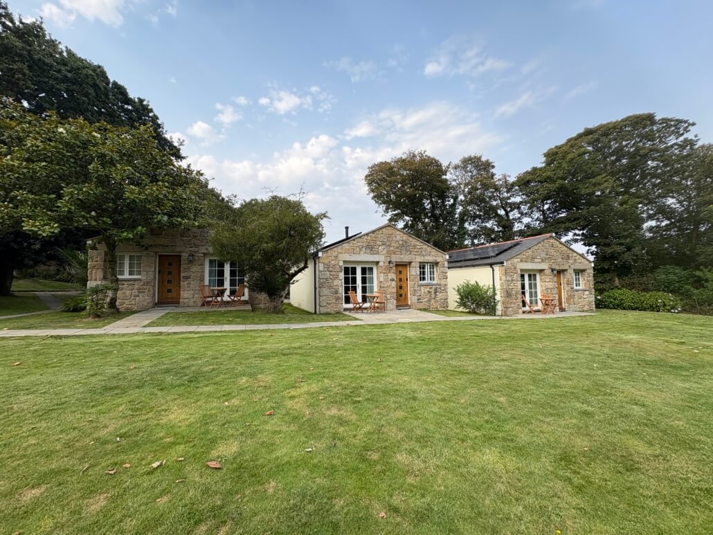 Three small stone cottages with wooden doors and outdoor seating are surrounded by grass and trees under a partly cloudy sky—perfect for a relaxing last minute Penzance getaway. Sancreed House Retreat