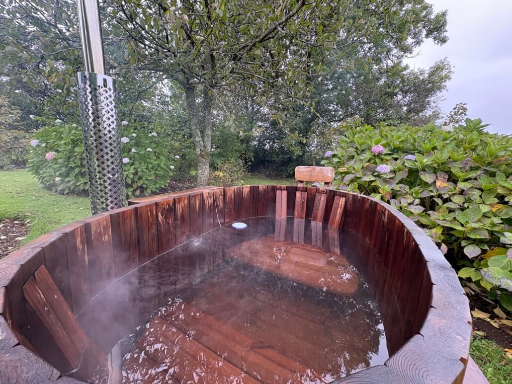 A steaming wooden hot tub filled with water sits outdoors on grass, surrounded by lush green bushes and trees under an overcast sky—perfect for a relaxing last minute Penzance escape. A metal stove is attached to the tub for heating. Sancreed House Retreat