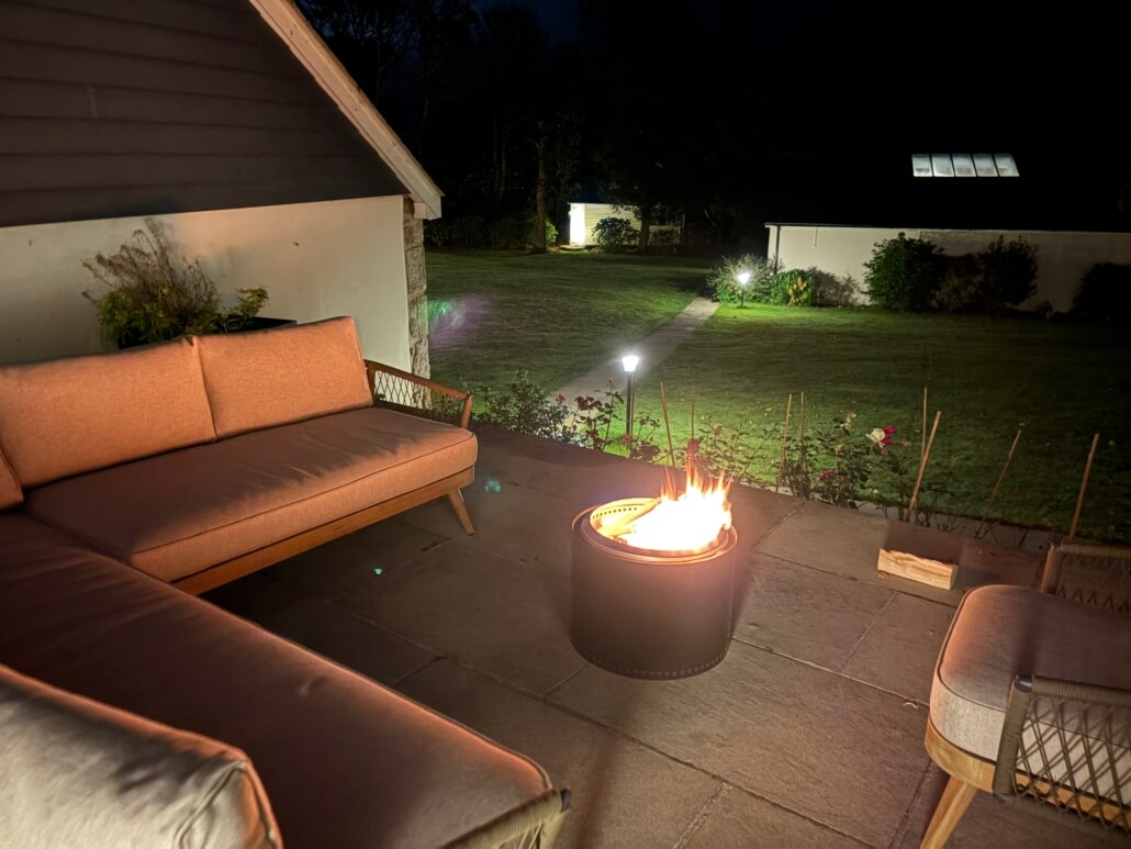 A cozy outdoor patio at night features a fire pit with bright flames, surrounded by cushioned chairs and a sofa—perfect for enjoying Last Minute Penzance getaways—overlooking a grassy yard with garden lights and distant buildings. Sancreed House Retreat