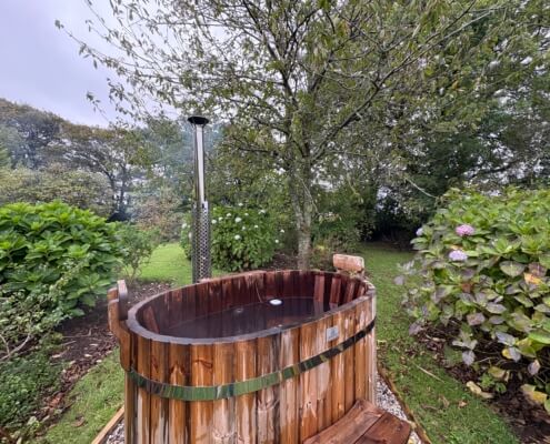 A wooden outdoor hot tub filled with water sits in a lush garden, surrounded by green shrubs, flowering bushes, and trees on a cloudy day. Steam rises from the tub, inviting you to enjoy warm water and explore additional services for ultimate relaxation. Sancreed House Retreat