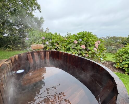 A steaming wooden hot tub sits in a garden surrounded by green bushes and blooming hydrangeas under a cloudy sky. Enjoy this peaceful outdoor scene with Additional Services to enhance your experience. Sancreed House Retreat