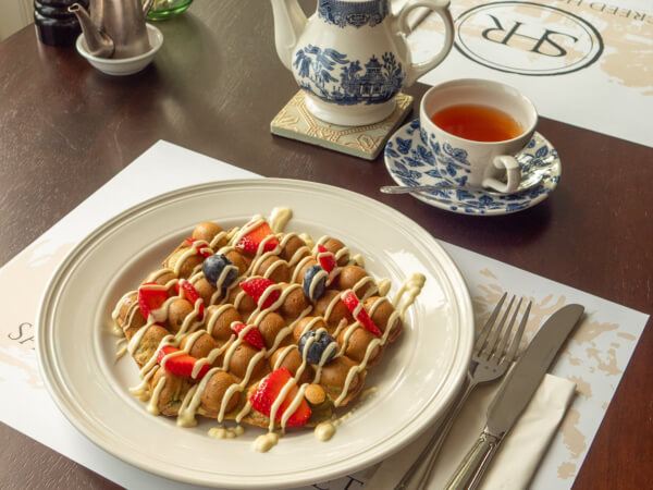 A plate of home-cooked waffles topped with strawberries, blueberries, and drizzled with cream sits next to a cup of tea in a blue-and-white cup and a matching teapot on a wholesome set dining table. Sancreed House Retreat