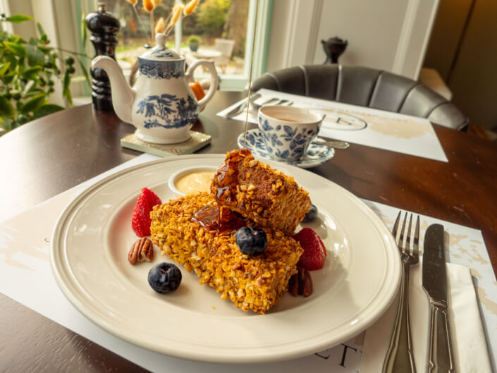 A plate of home-cooked, wholesome granola-crusted French toast topped with syrup, blueberries, strawberries, and pecans sits on a table; behind it are a teapot, teacup, and a window with greenery outside. Sancreed House Retreat