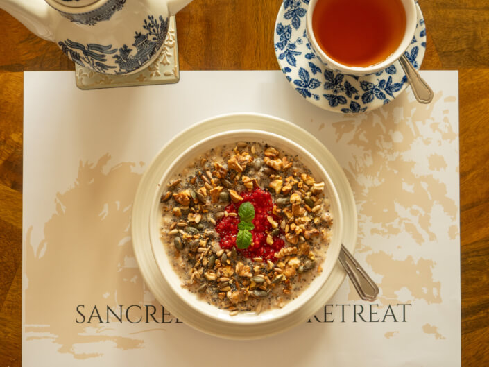 A bowl of home-cooked oatmeal topped with granola, seeds, and a raspberry garnish sits on a placemat next to a cup of tea and teapot in blue and white china on a wooden table, creating a wholesome breakfast scene. Sancreed House Retreat