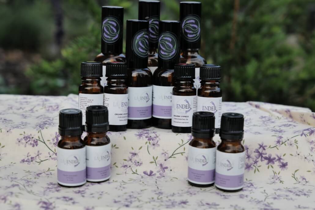 Several small amber bottles of essential oils with lavender-themed labels are arranged on a floral-patterned cloth outdoors, with greenery in the blurred background. Sancreed House Retreat
