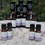 Several small amber bottles of essential oils with lavender-themed labels are arranged on a floral-patterned cloth outdoors, with greenery in the blurred background. Sancreed House Retreat