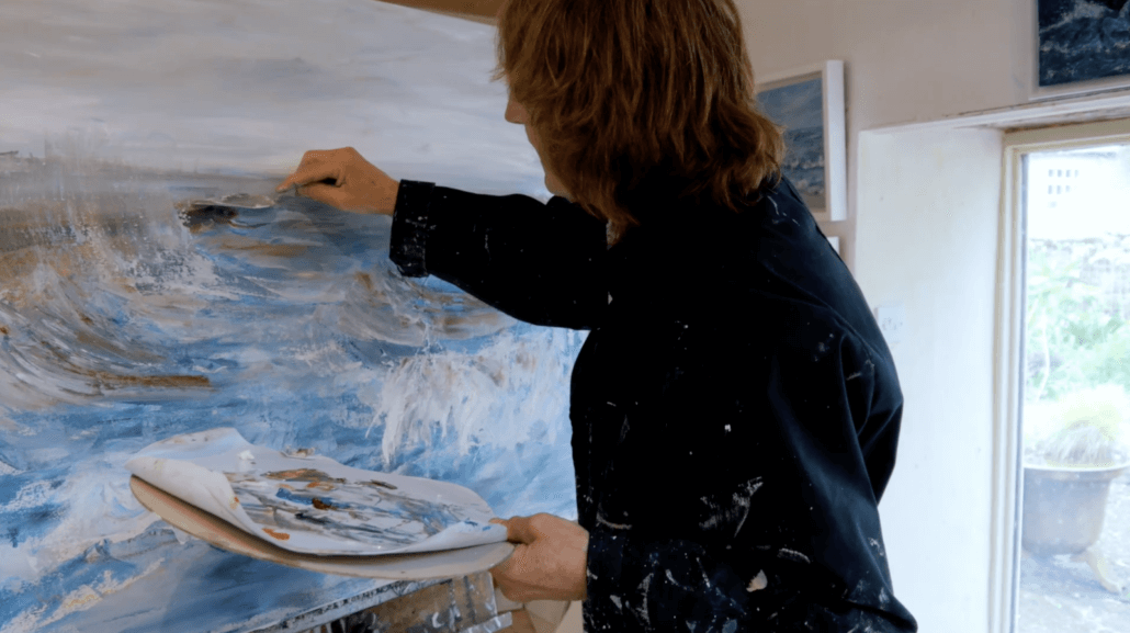 A person with shoulder-length hair wearing a dark, paint-splattered shirt paints an abstract seascape on a canvas. They hold a palette with mixed colors and stand in a bright art studio. Sancreed House Retreat