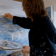 A person with shoulder-length hair wearing a dark, paint-splattered shirt paints an abstract seascape on a canvas. They hold a palette with mixed colors and stand in a bright art studio. Sancreed House Retreat